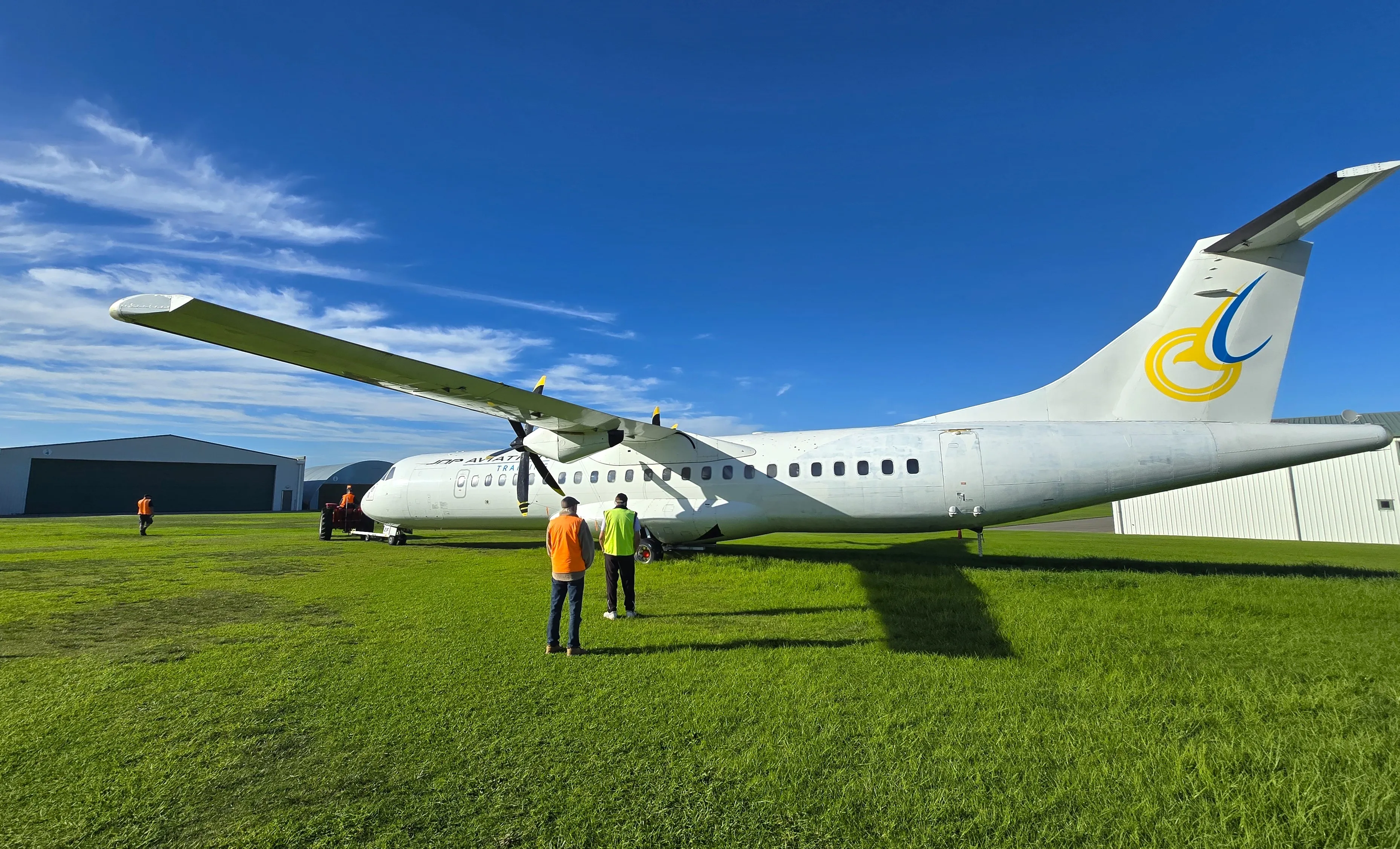 Side view of a white ATR airplane on green grass with three workers in high-visibility vests nearby under a blue sky.