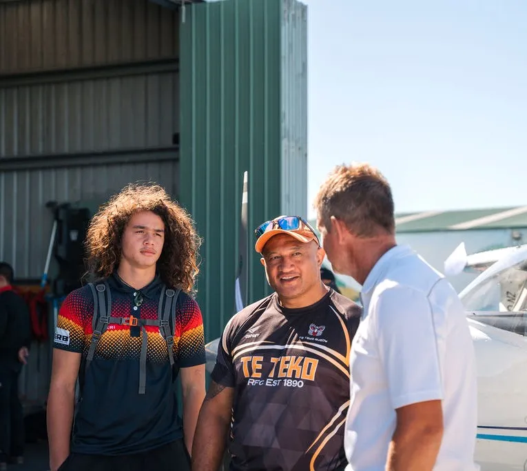 Three men standing outdoors near an airplane hangar having a conversation, one wearing a colorful shirt, one in a Te Teko rugby shirt, and one in a white shirt.