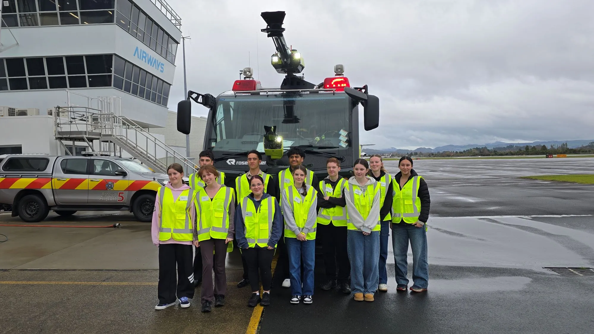 Group of nine people wearing yellow safety vests standing in front of a fire truck and an airport vehicle on a wet tarmac.