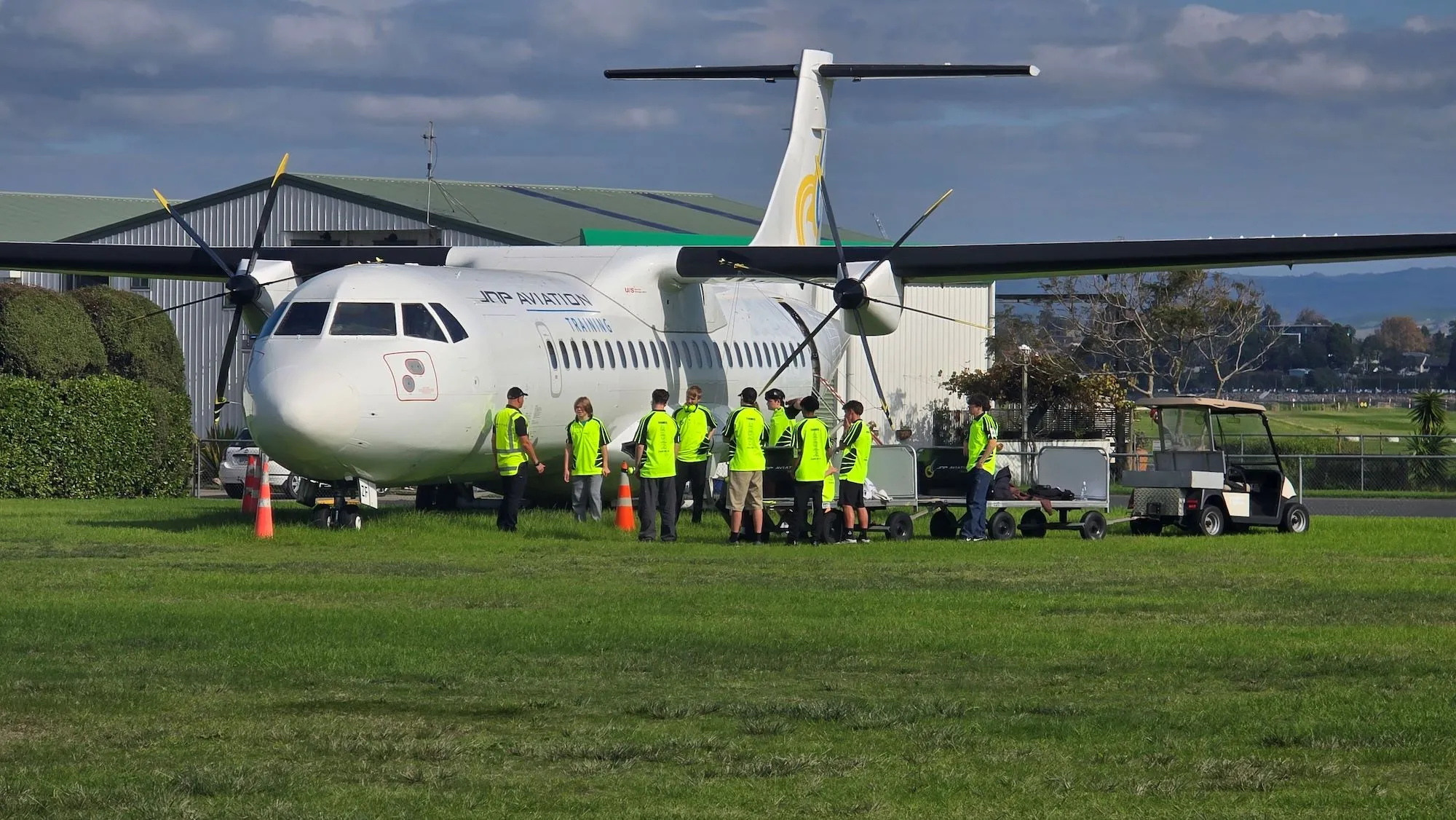 Group of people wearing neon yellow safety vests standing near a white training aircraft on a grassy airfield.
