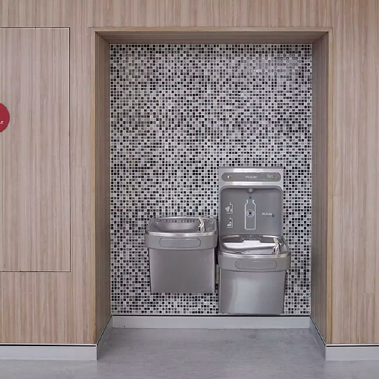 Stainless steel drinking fountains installed in a wall recess with small black and white mosaic tiles and wood paneling on sides.