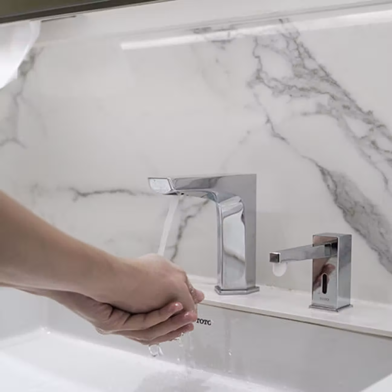 Hands under running water from a modern chrome bathroom faucet with marble wall background.