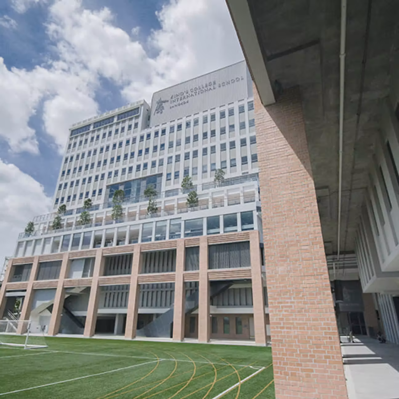Modern multi-story school building with large windows, brick pillars, and a green sports field in front under a partly cloudy sky.
