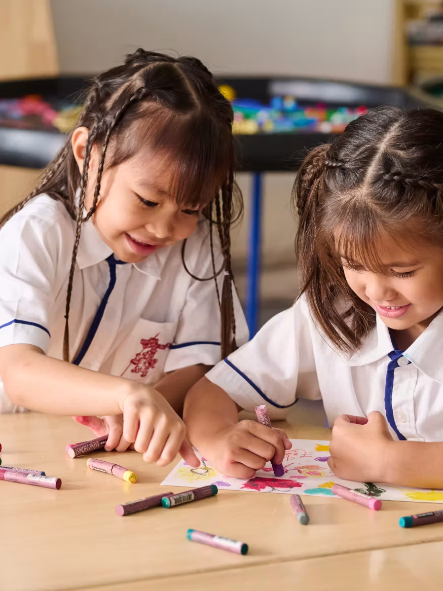 Two girls in school uniforms drawing with crayons on paper at a table, smiling and focused.