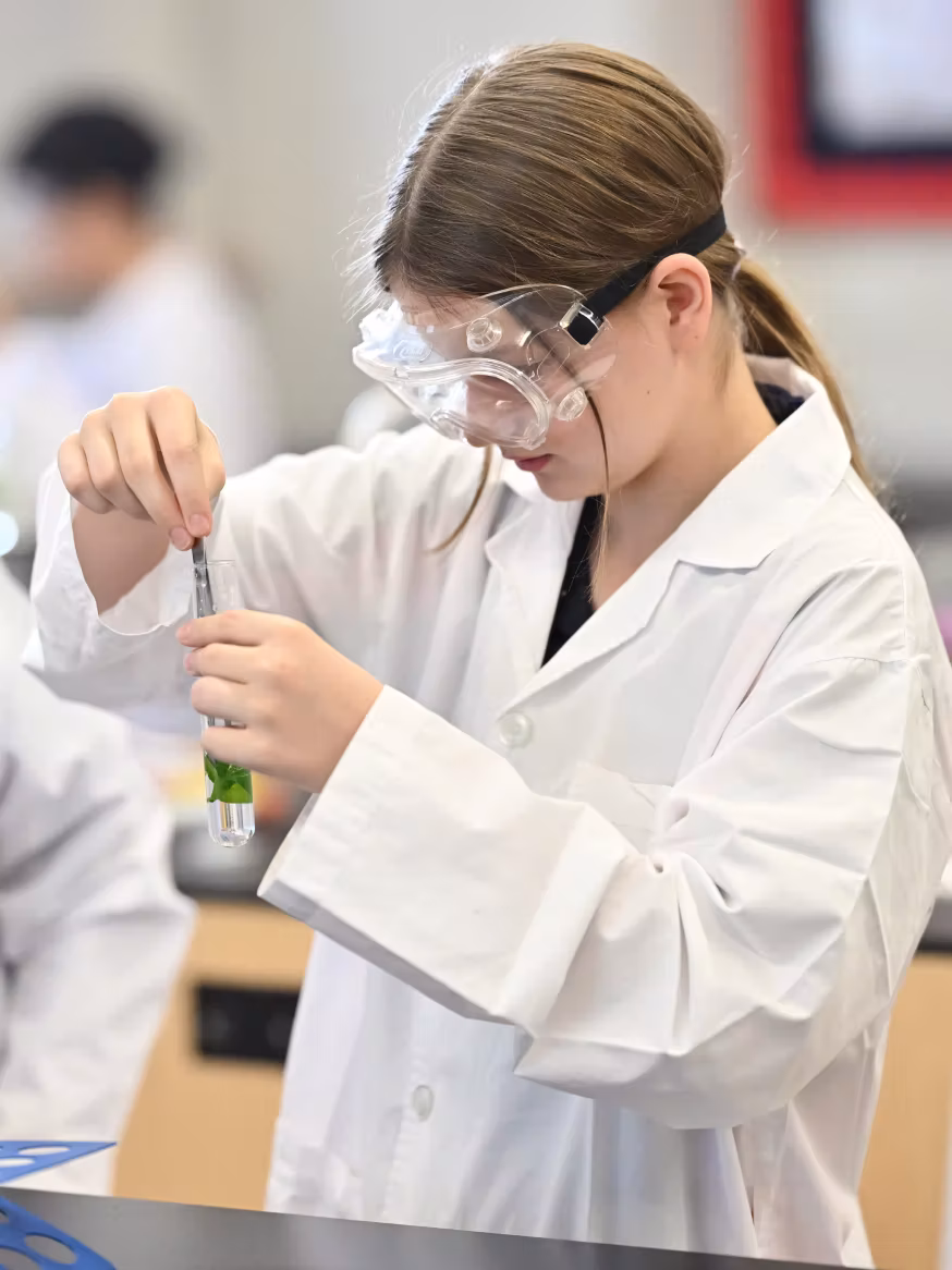 Young girl in safety goggles and lab coat stirring liquid with leaves in a test tube in a lab.