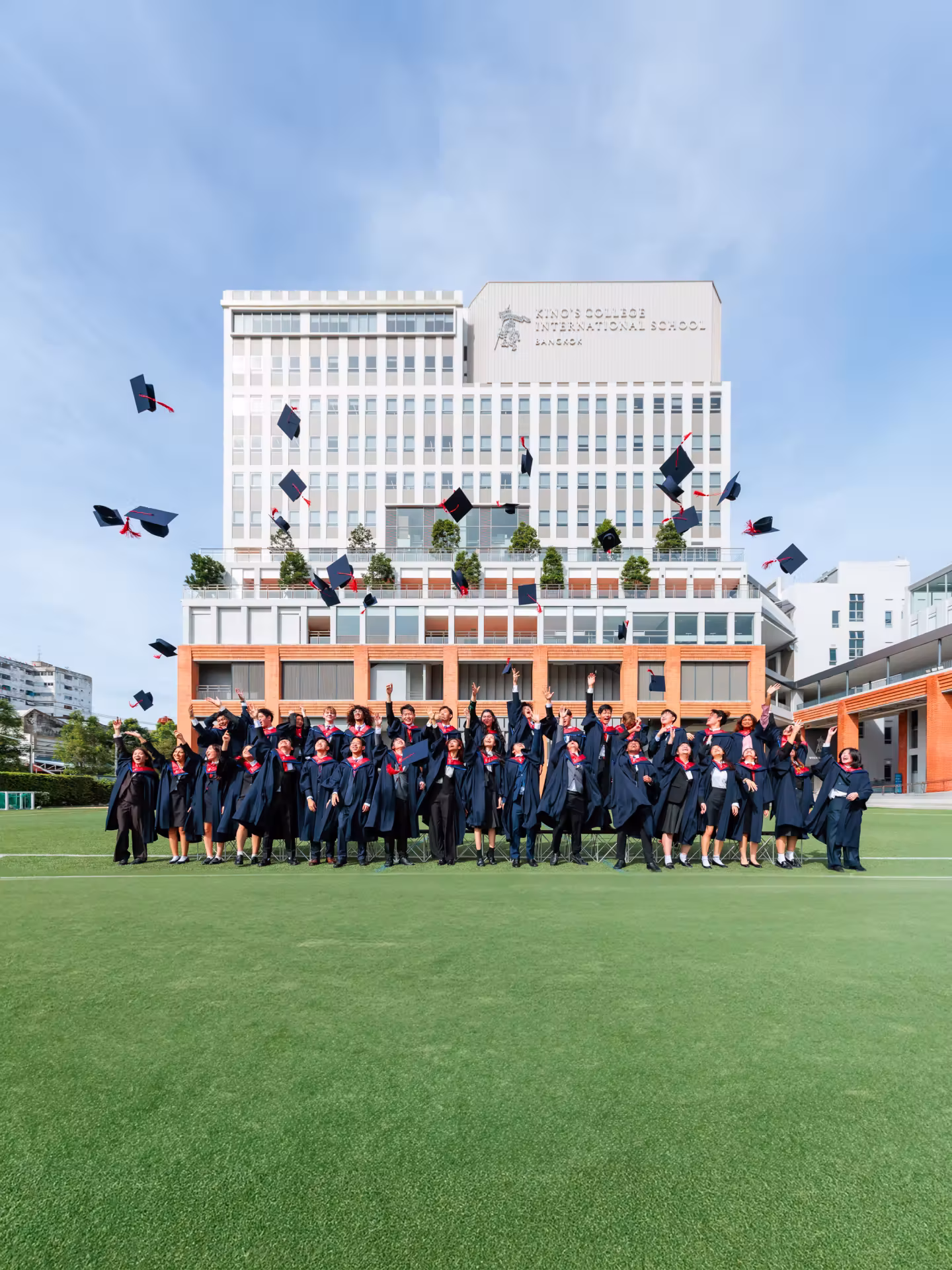 Group of graduates in blue gowns and red stoles throwing caps into the air in front of a modern school building.