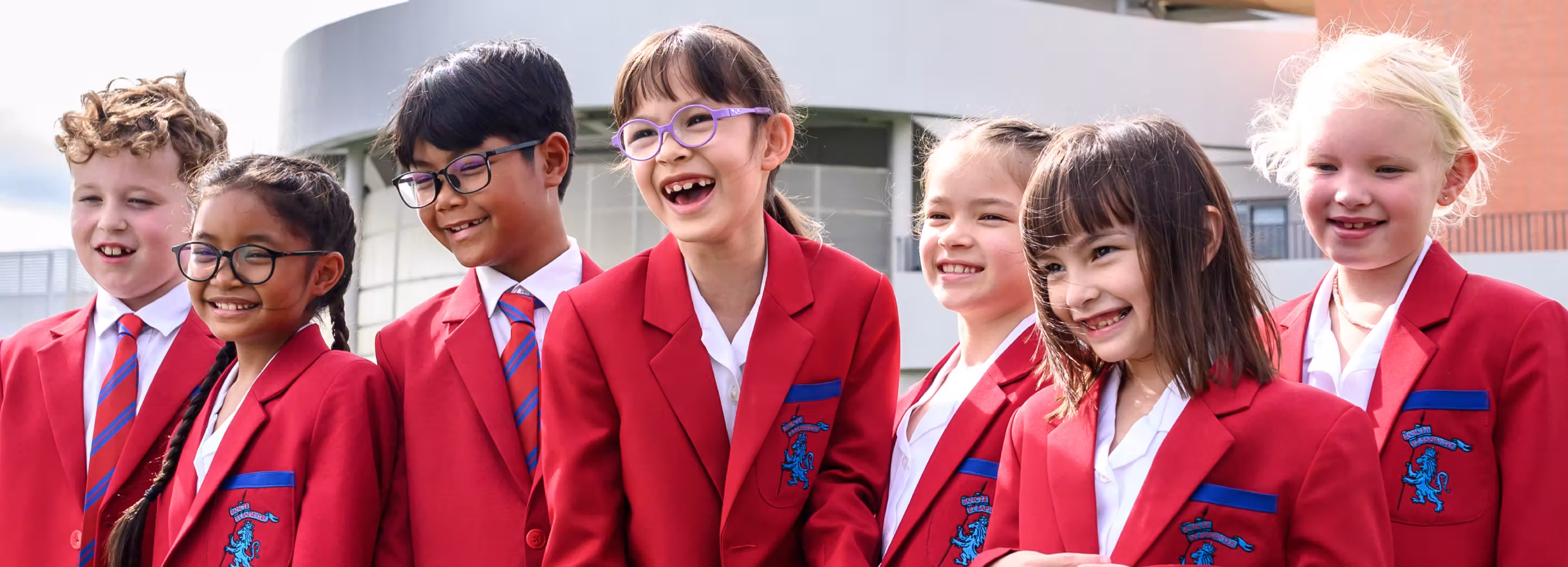Group of seven happy primary school children wearing red blazers and striped ties outdoors.