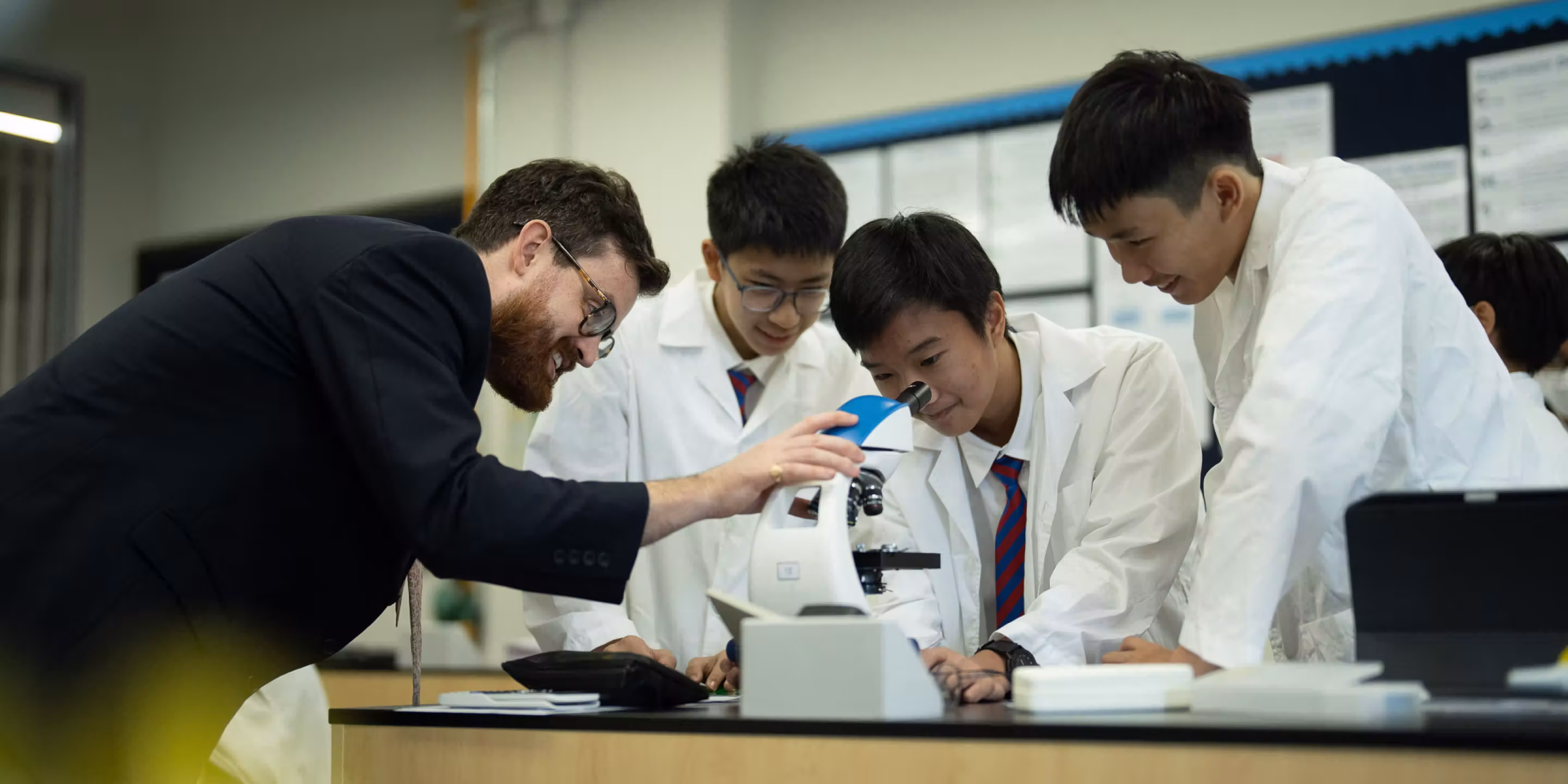 Teacher guiding students in lab coats as one looks through a microscope in a science classroom.