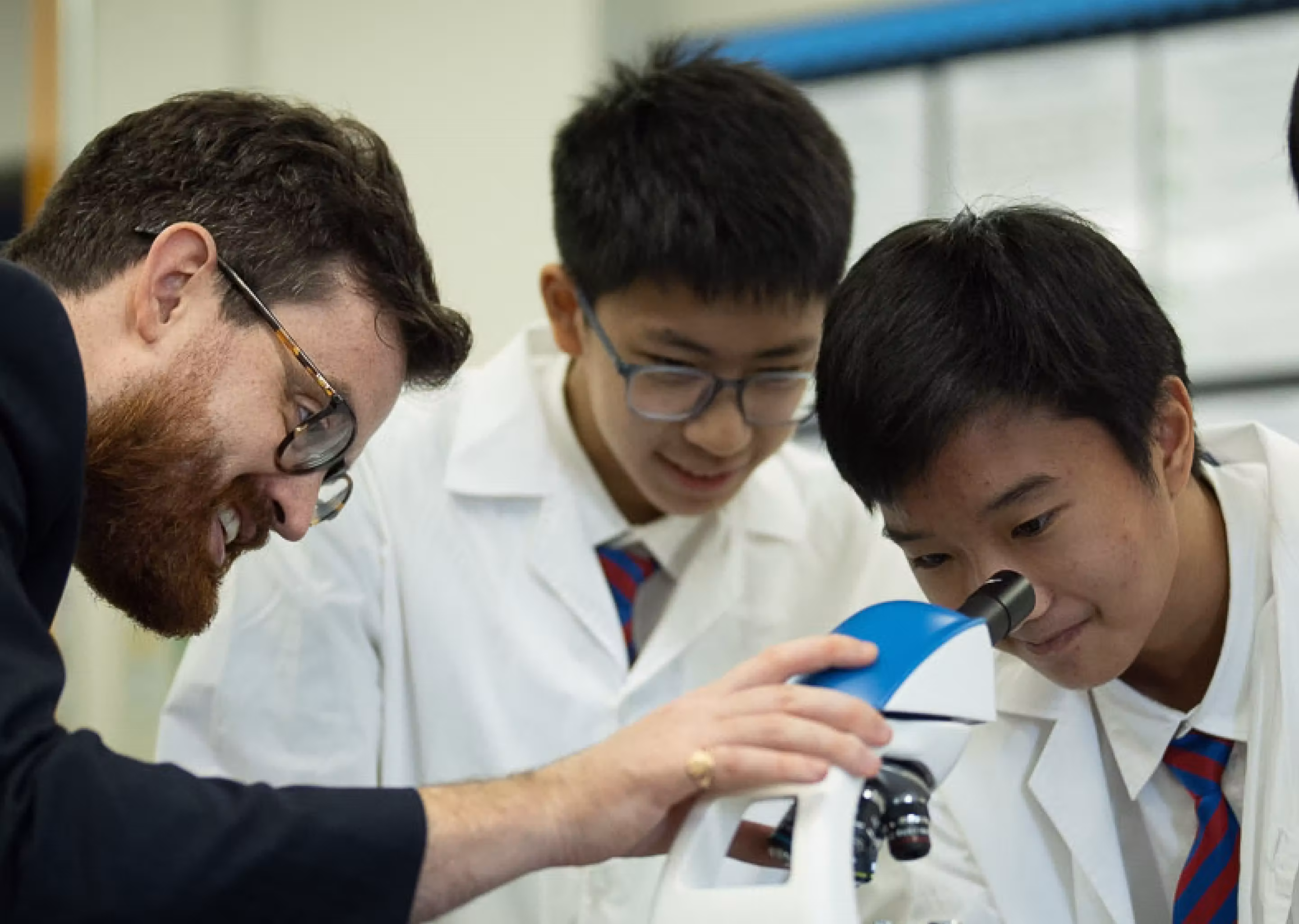 Teacher and two students in white coats examine a specimen using a microscope in a classroom.