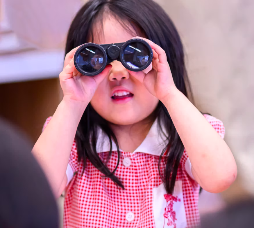 Young girl in red check dress looking through black binoculars indoors.