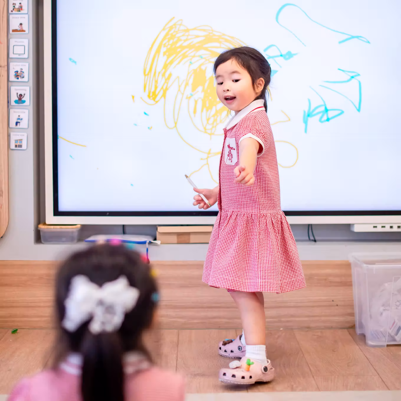 A young girl in a red dress pointing while standing in front of a whiteboard with colorful scribbles.