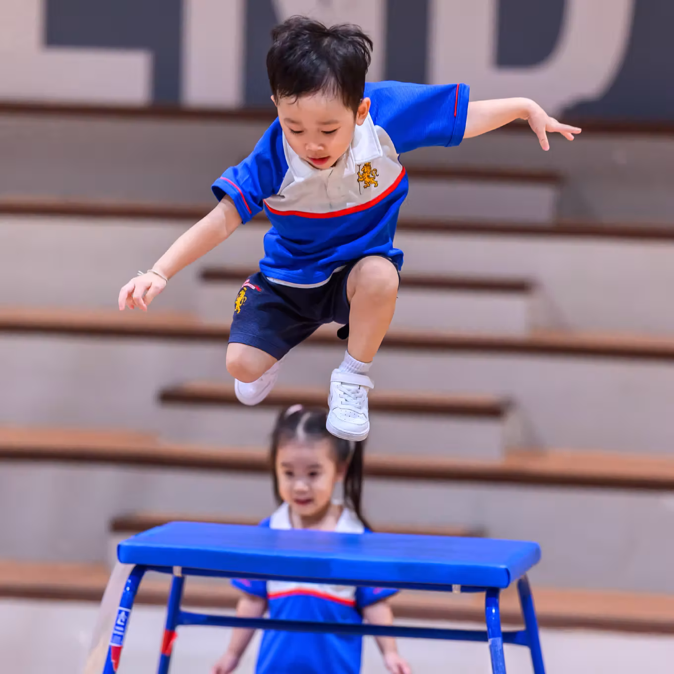Young boy in blue sportswear jumping over a blue gym bench with a girl watching behind him.