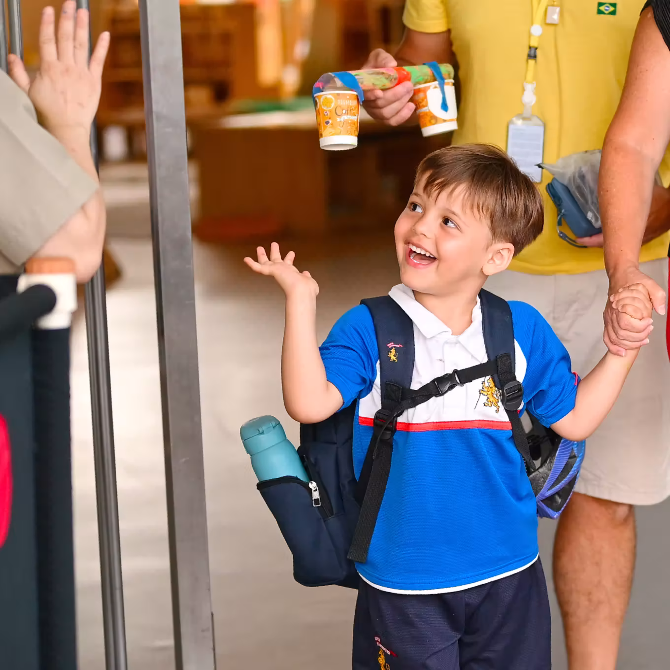 Smiling boy with backpack and water bottle holding hands with adult entering building greeted by raised hand.