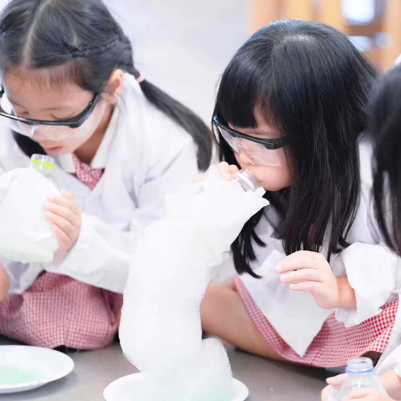Two girls in goggles and lab coats blowing into bottles creating foam in a science experiment.
