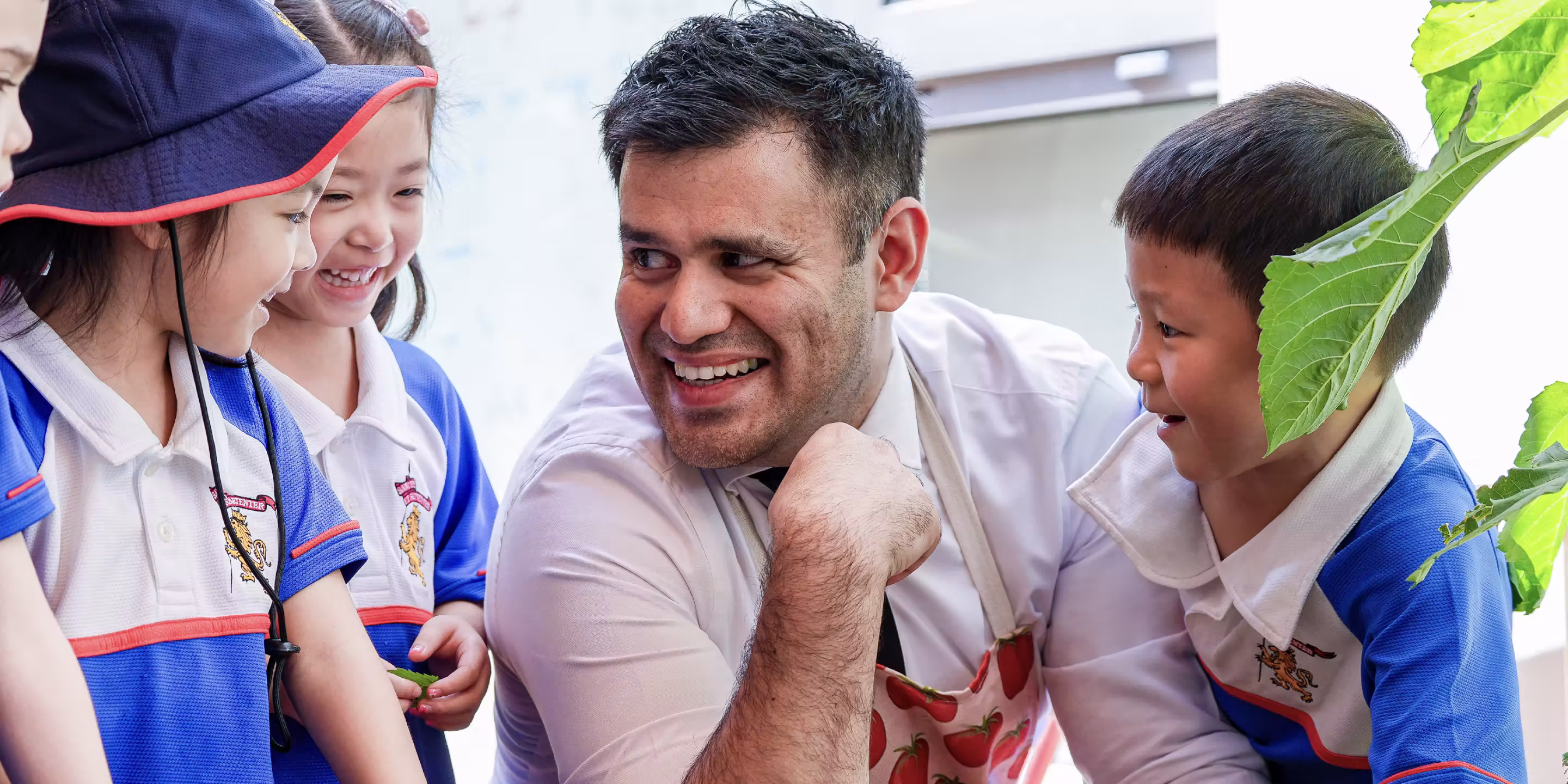 Smiling adult with apron engaging happily with four young children in school uniforms.