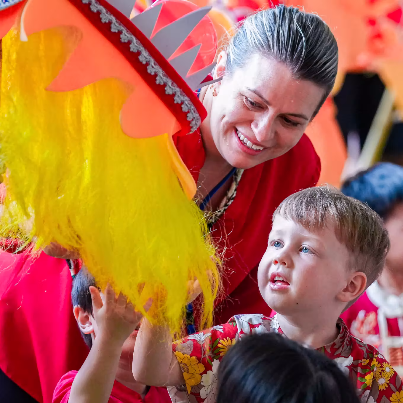 Smiling woman helps young boy touch bright yellow puppet during colorful activity.