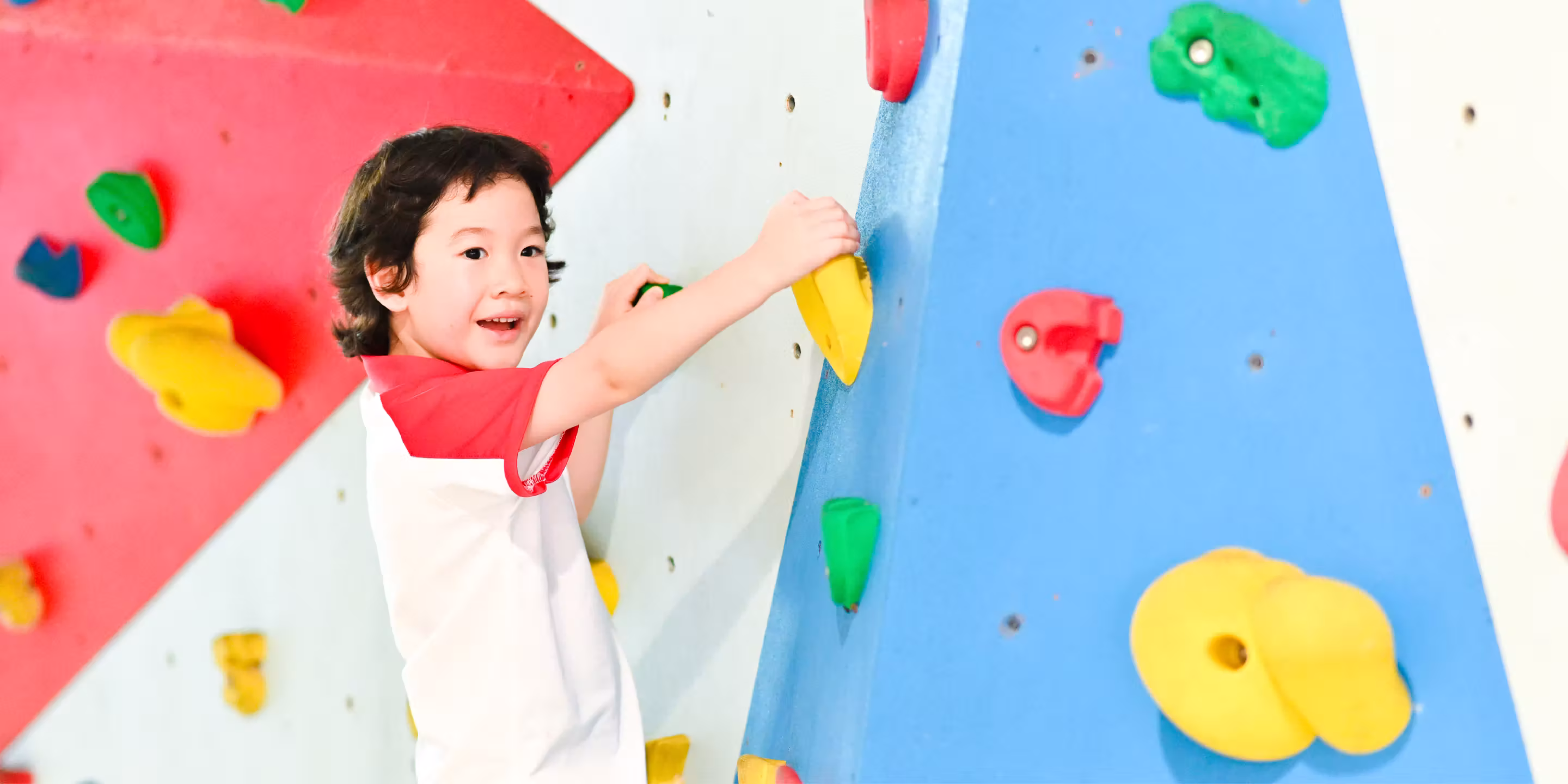 Child smiling while climbing a colourful indoor rock climbing wall with red, blue, yellow, and green holds.