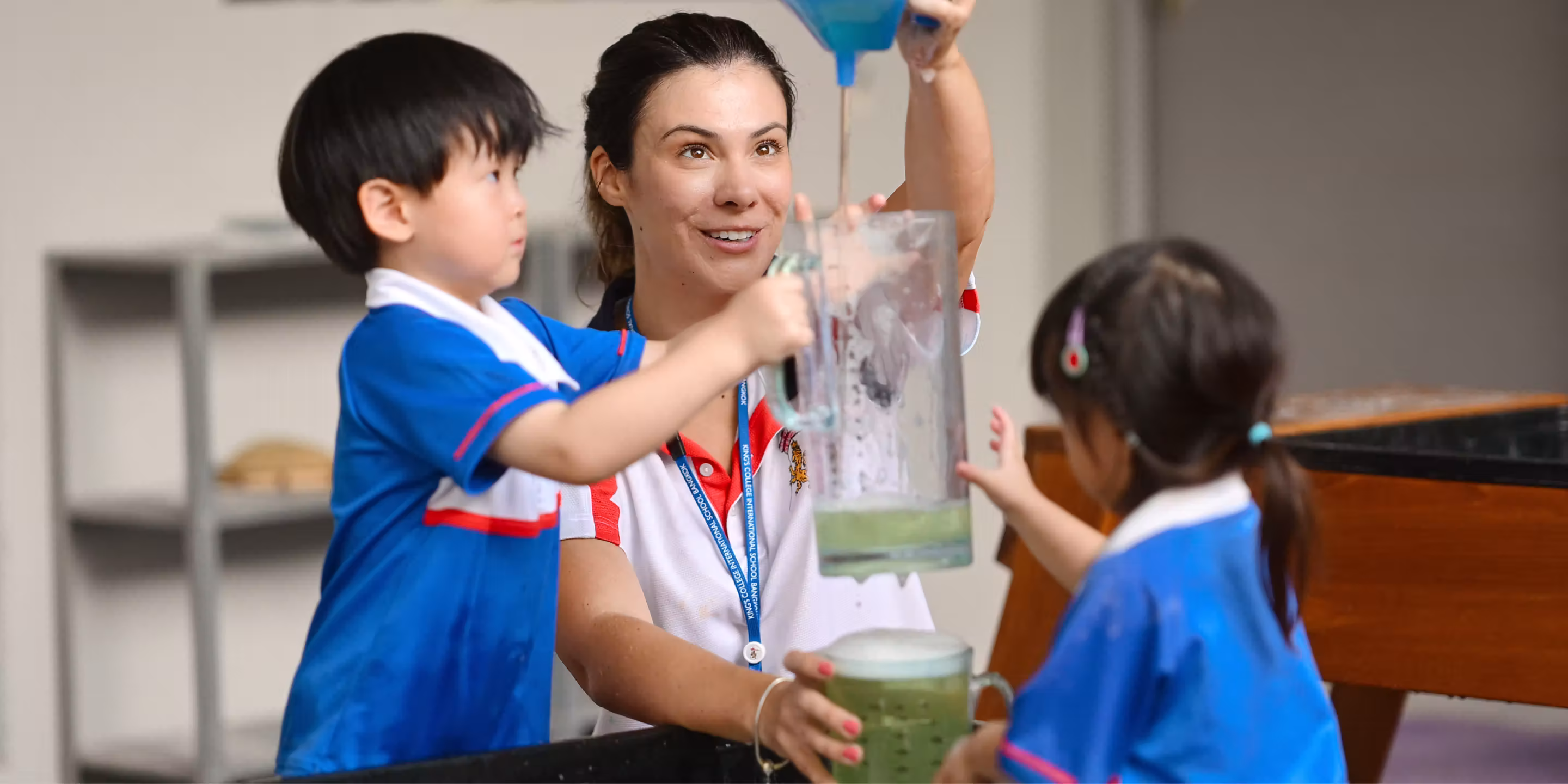 Teacher and two children in blue uniforms doing a water experiment with jugs in a classroom.