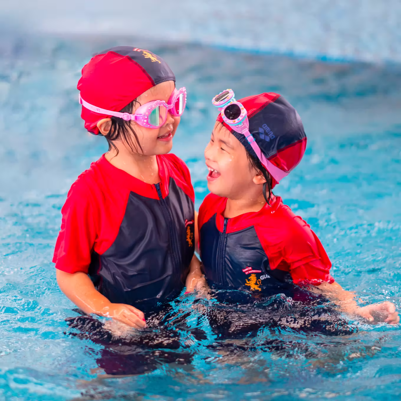 Two children in red and black swim gear and goggles playing and smiling in a swimming pool.