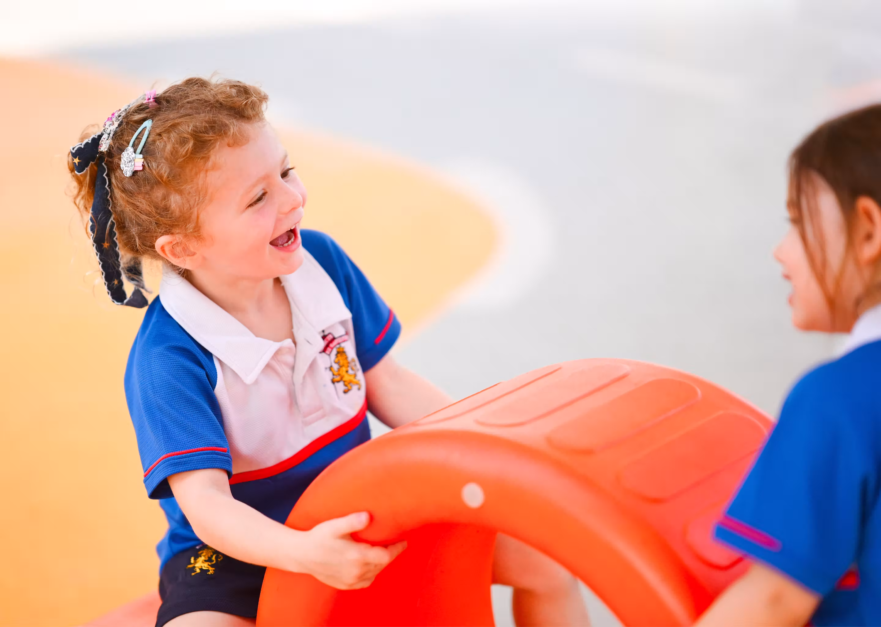 Two young girls smiling and playing on an orange playground toy outdoors.