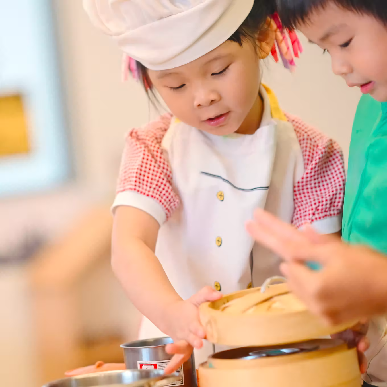 Young girl wearing a chef’s hat and apron lifting bamboo steamer lid while a boy assists her.