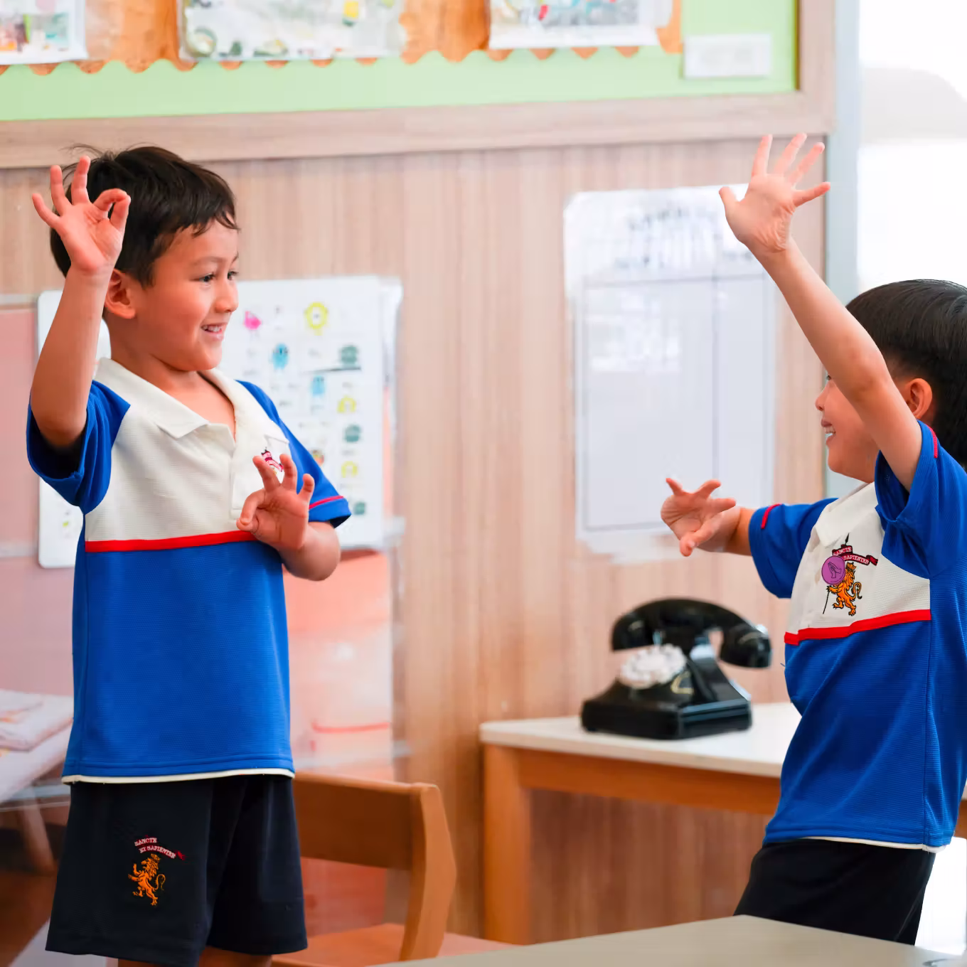 Two boys in blue school uniforms playfully raising their hands inside a classroom.