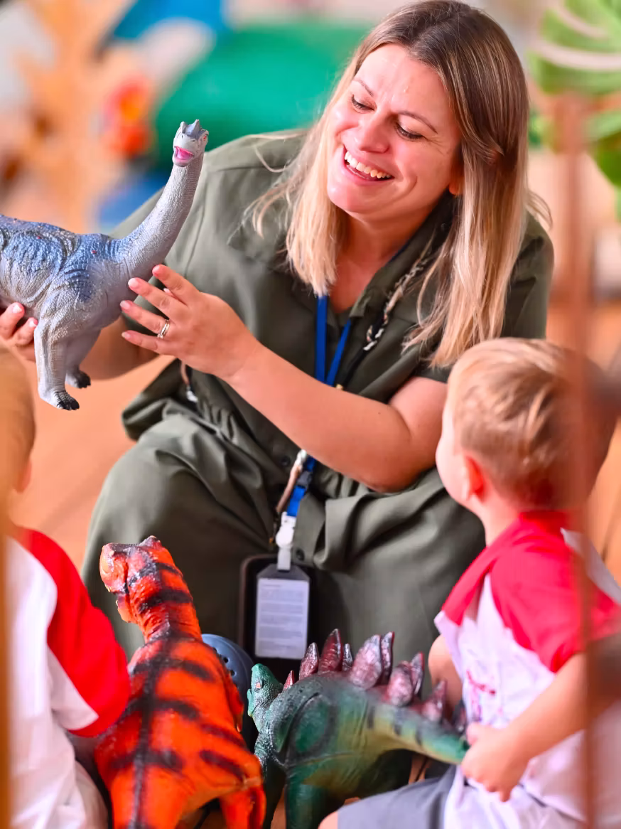 Smiling woman shows children a toy dinosaur during a play activity with dinosaur figures.