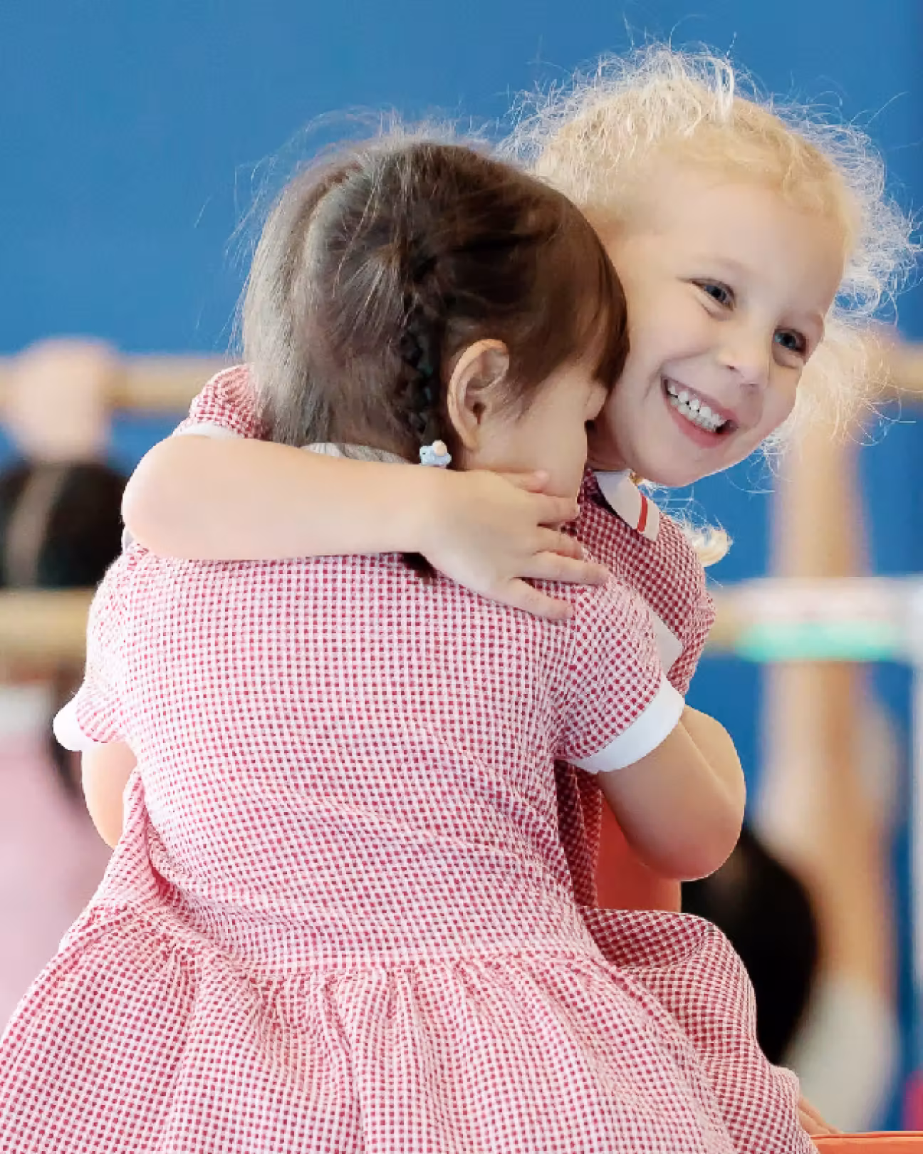 Two young girls wearing red checkered dresses hugging and smiling indoors with a blue background.