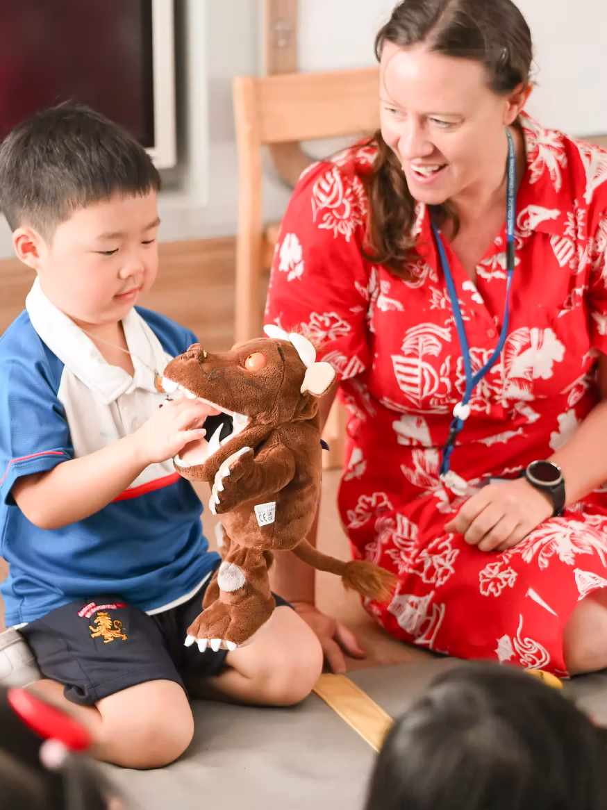 Child playing with a brown stuffed animal while a woman in a red dress watches and smiles nearby.