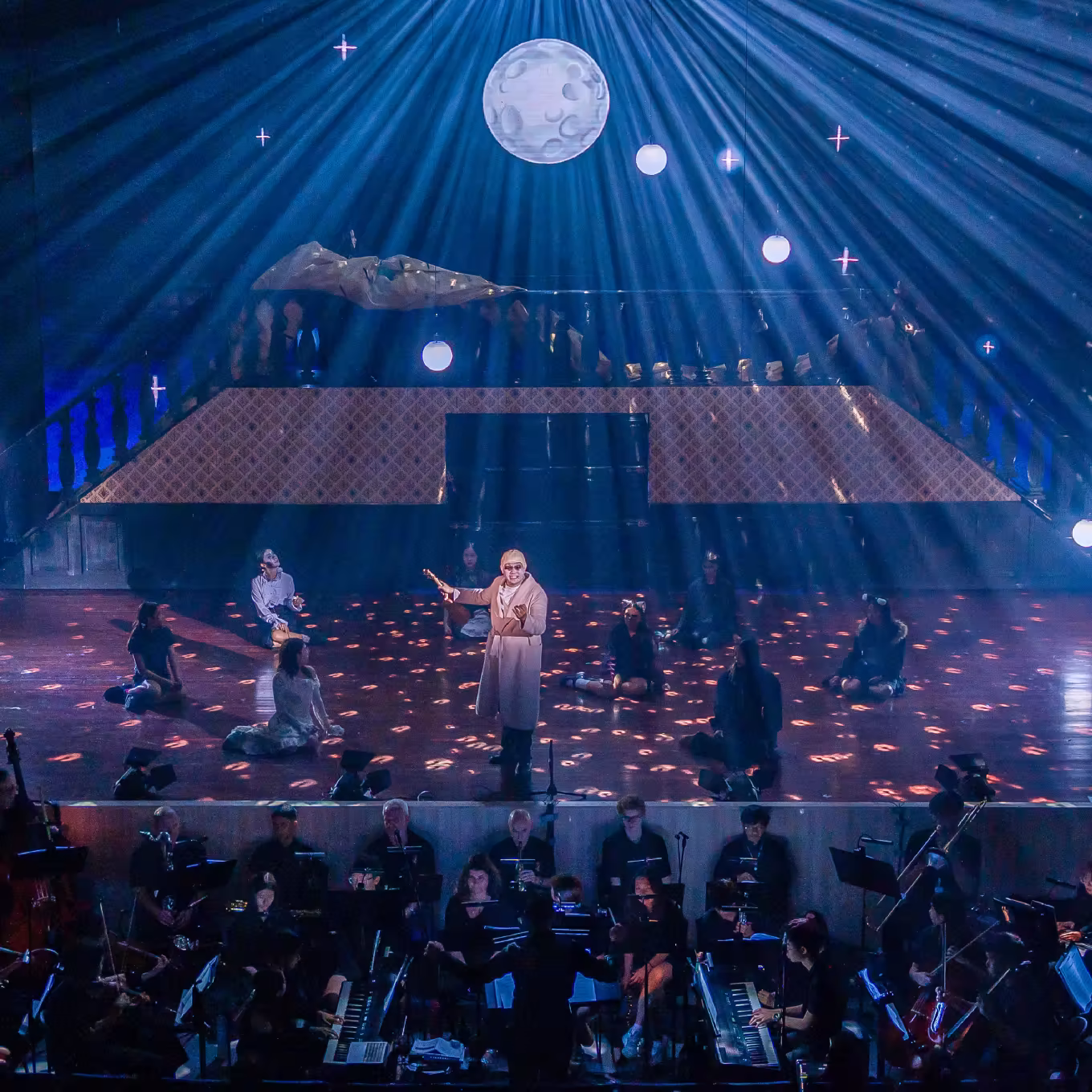 Theatrical stage with actors sitting and standing under moonlight effects and orchestra playing below.