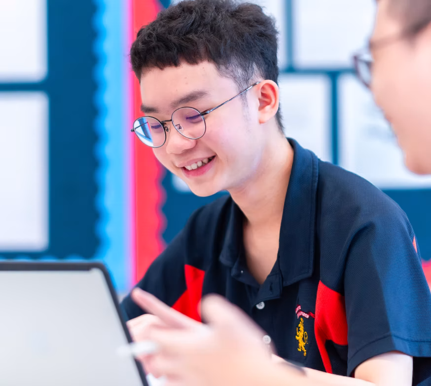 Smiling student wearing glasses and school uniform using a laptop in a classroom setting.