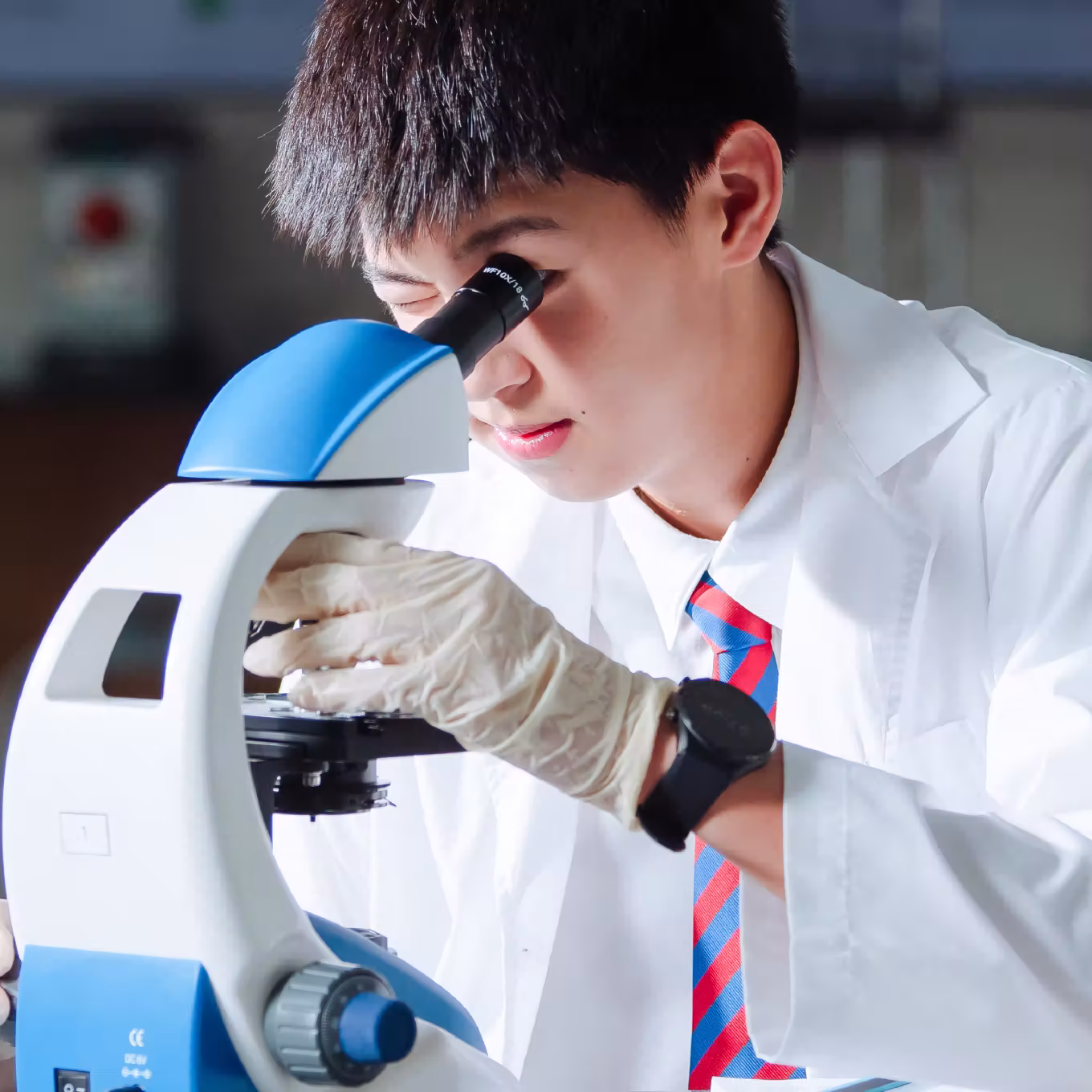 Student in lab coat and gloves looking into a microscope in a laboratory setting.