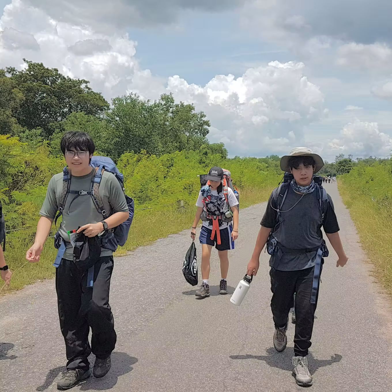 Three hikers walking on a road carrying backpacks and supplies under a cloudy sky.