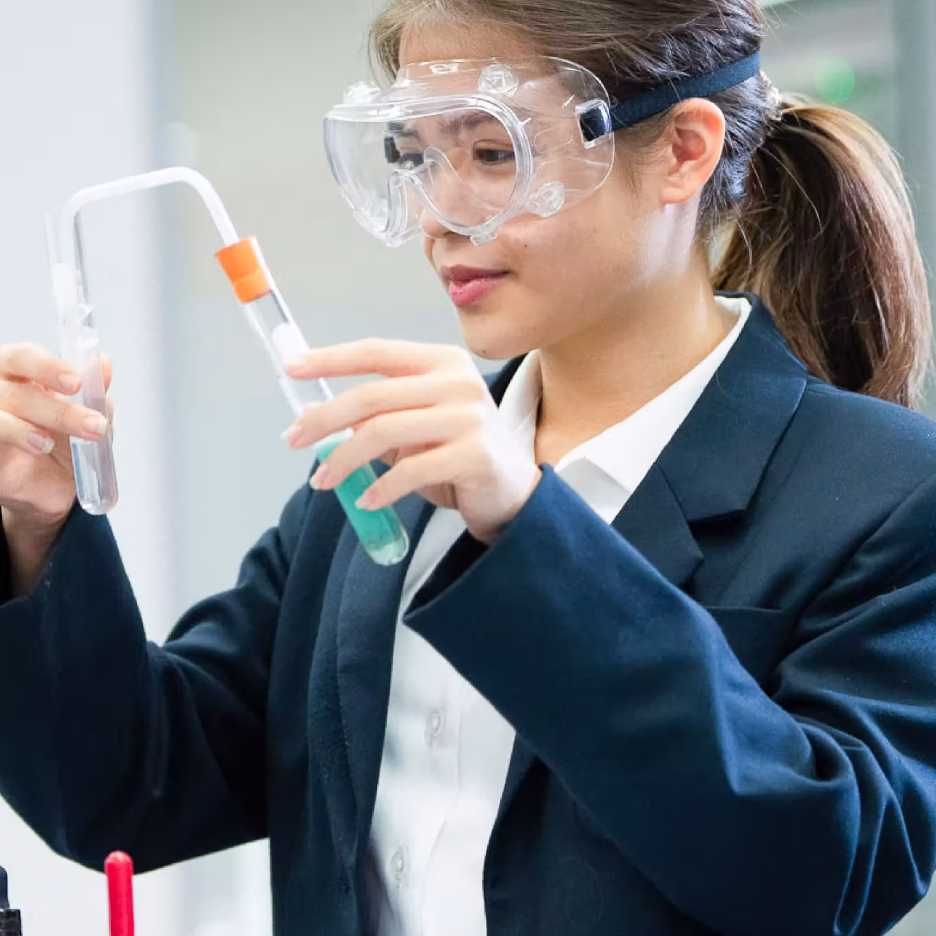 Young woman wearing safety goggles handling test tubes with liquids in a science lab.