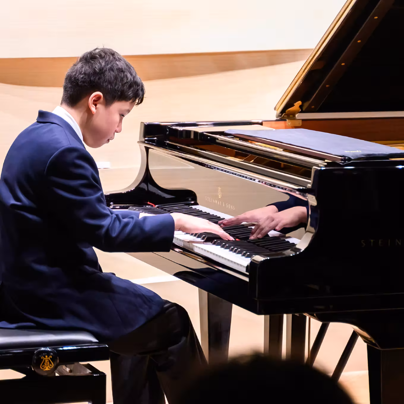 Young boy in suit playing a grand piano, focused on the keys in a well-lit room.