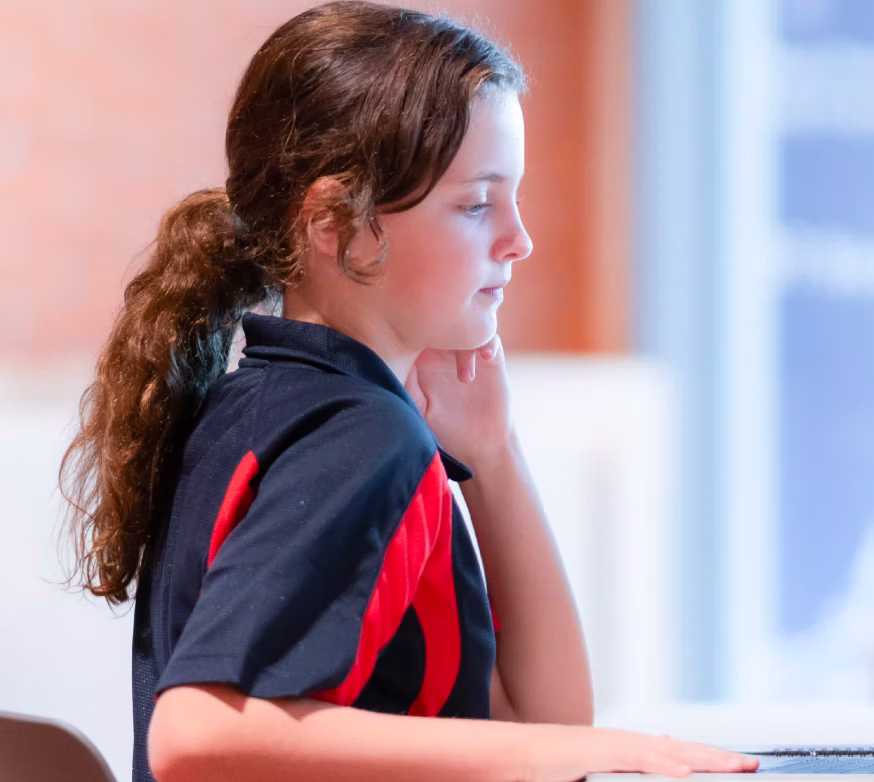 Young girl with curly hair in a ponytail, wearing a black and red shirt, sitting and looking focused.