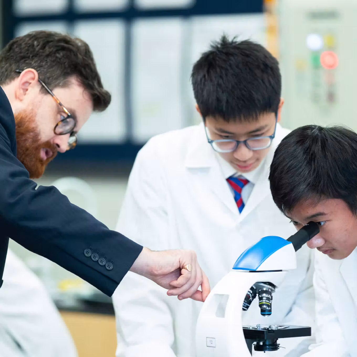 Teacher guiding two students in lab coats using a microscope in a science classroom.