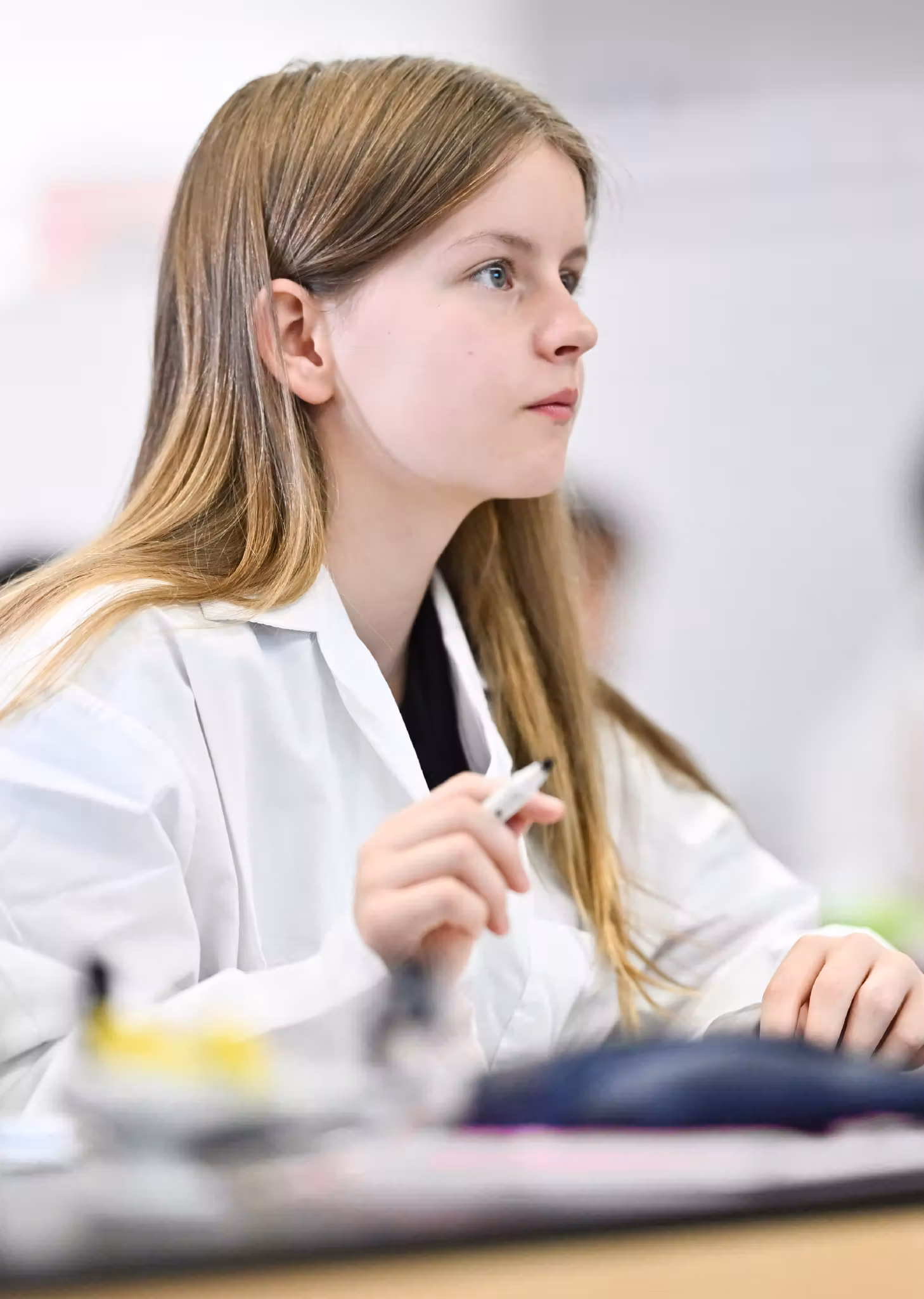Young girl with long blonde hair in white coat holding a pen, looking intently to the side.