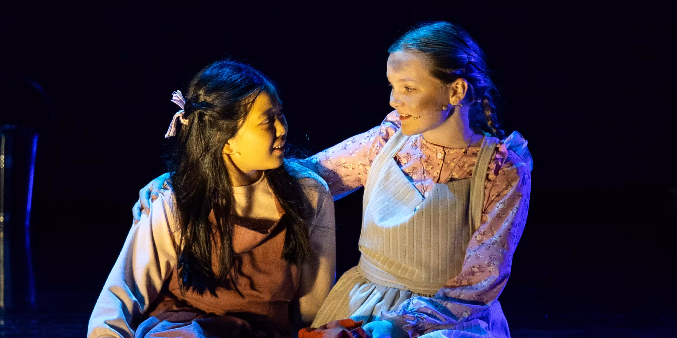 Two young women in vintage dresses perform an emotional scene on stage under blue lighting.