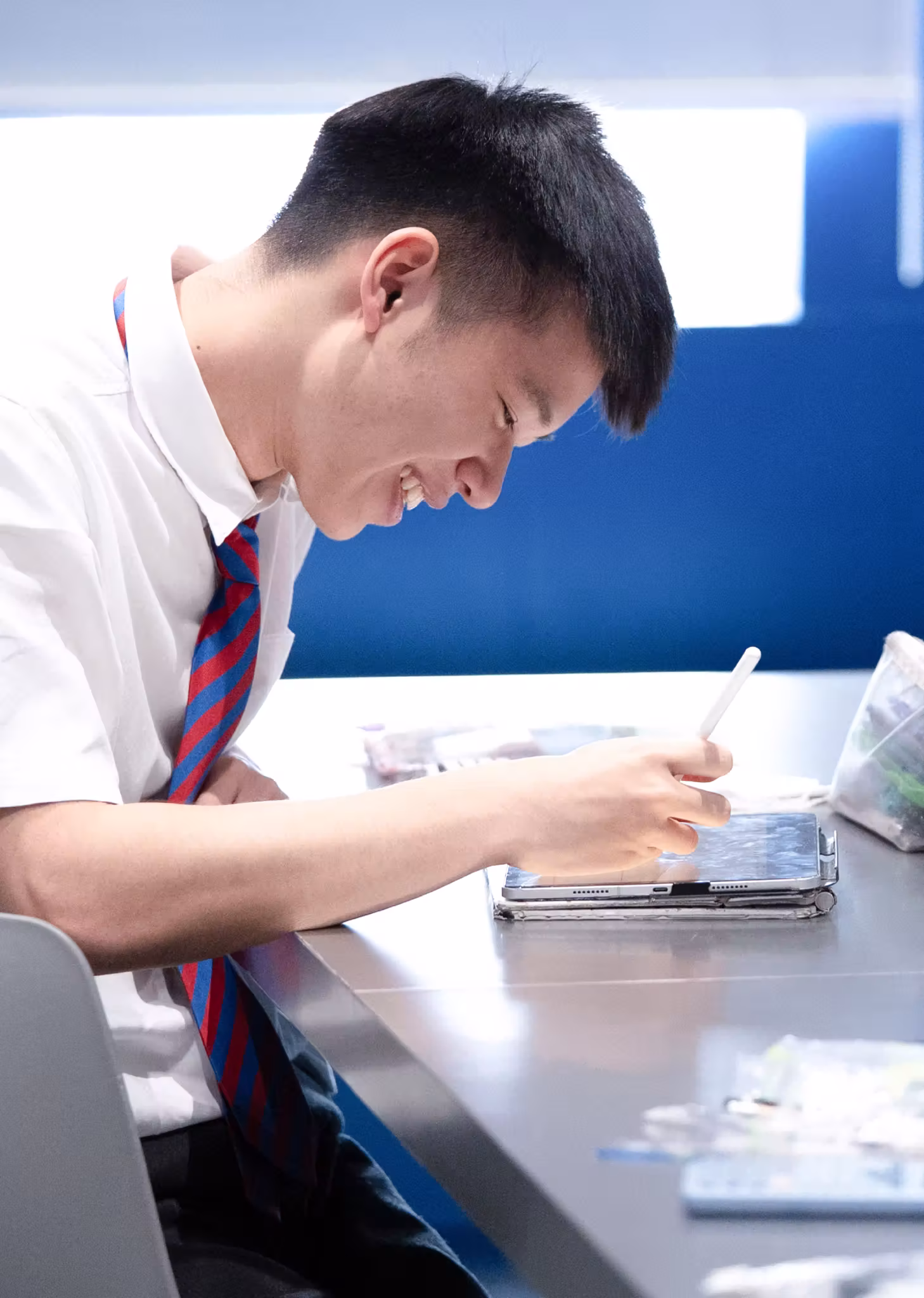 Student in white shirt and striped tie smiling while using a stylus on a tablet at a desk.