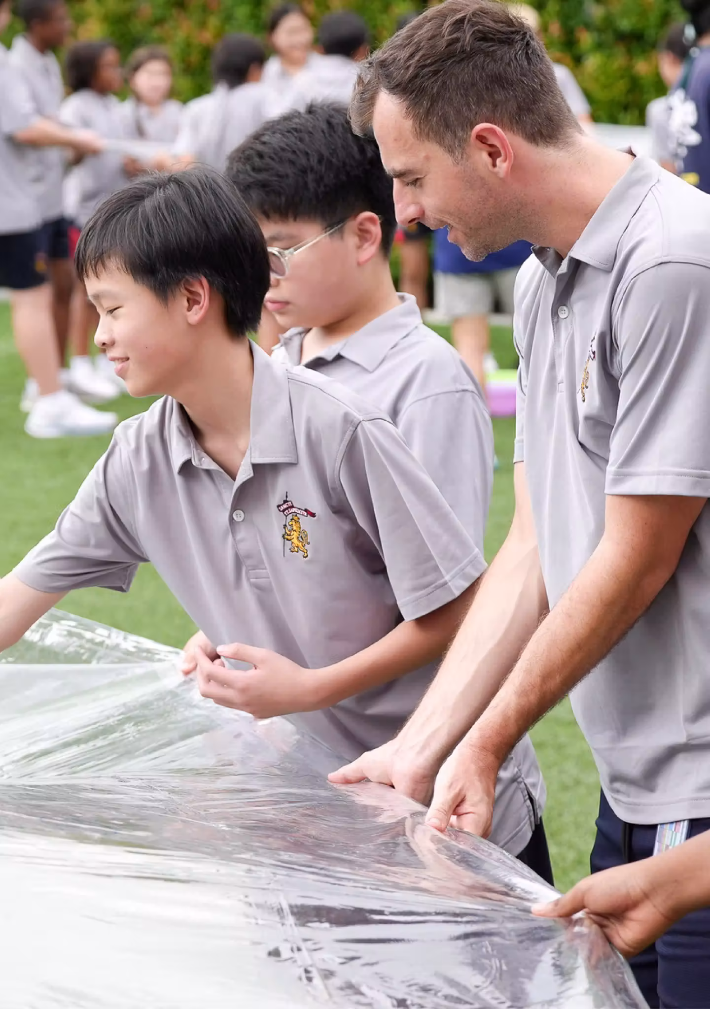 Adults and children in matching grey shirts handling a large clear plastic sheet outdoors.