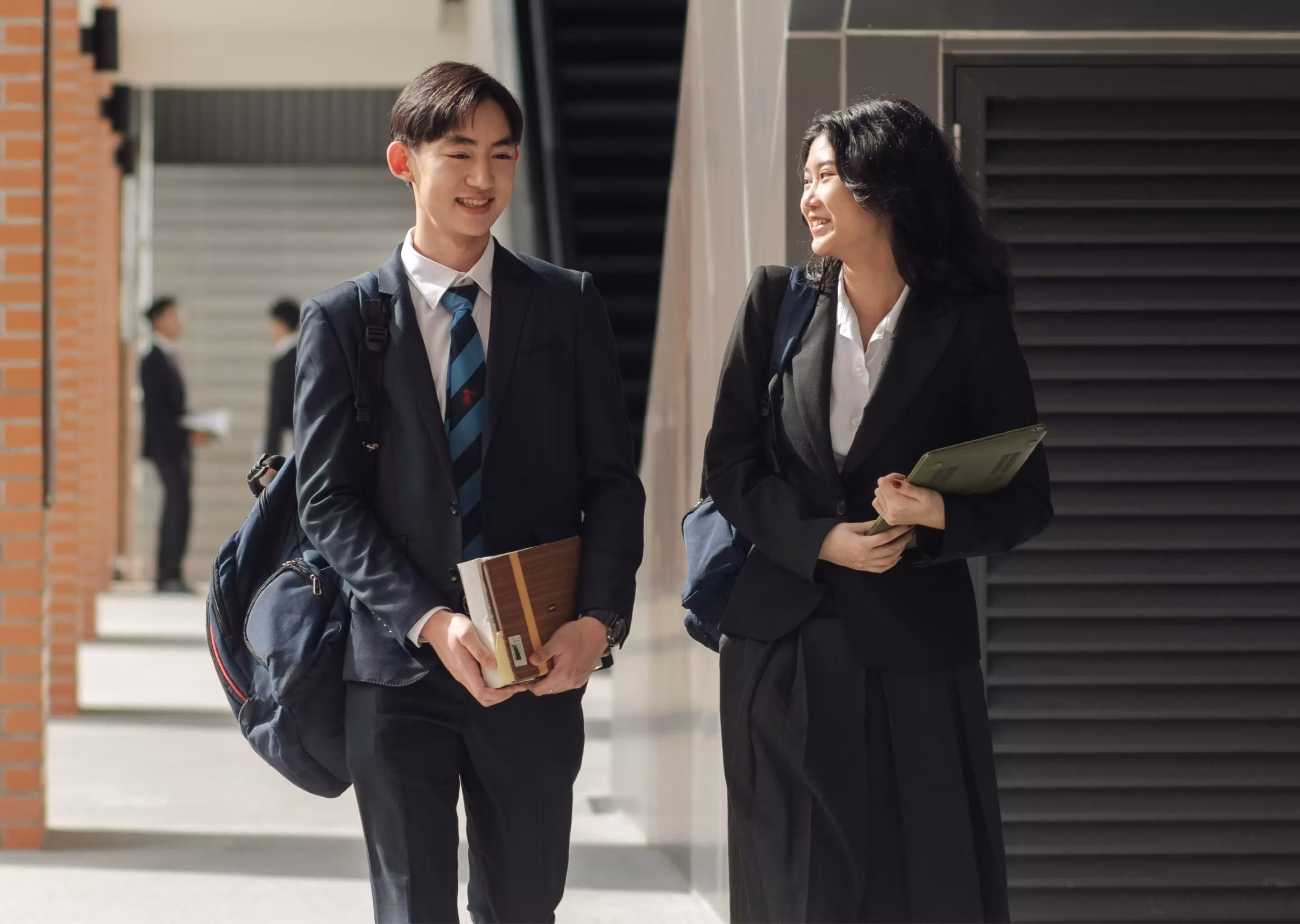 Two students in formal uniforms walking outdoors, smiling and holding books with backpacks on.