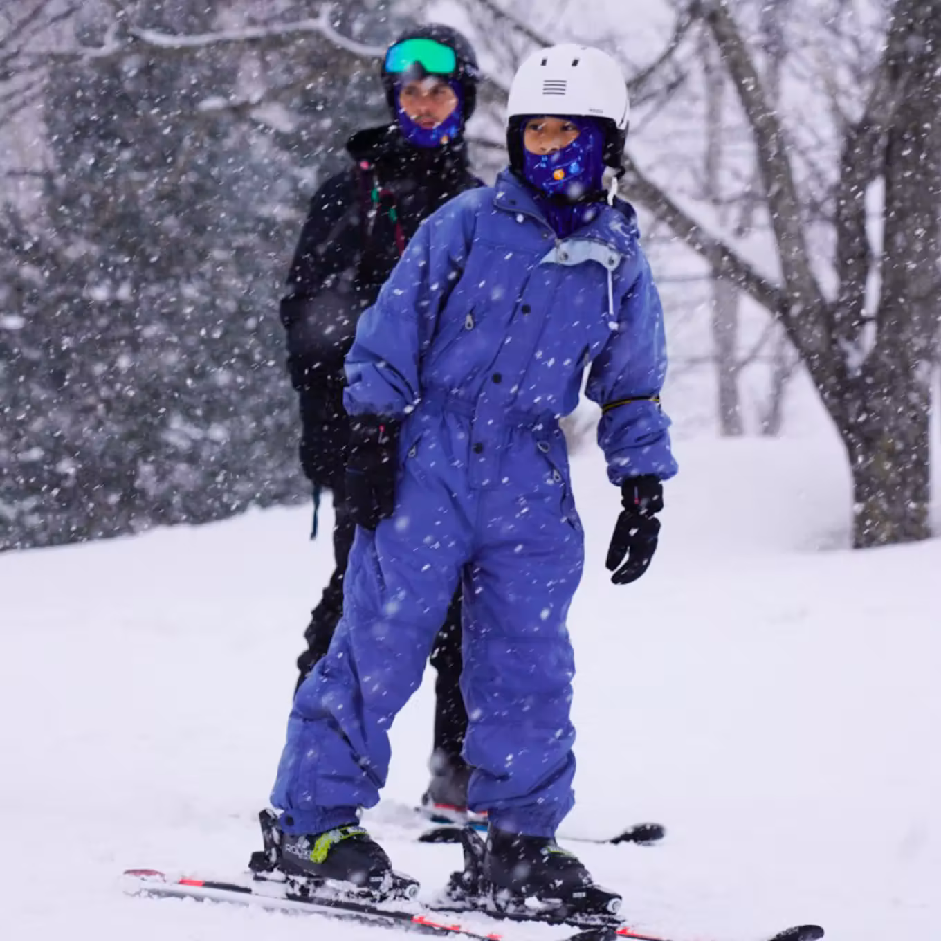 Child in blue snowsuit and white helmet skiing with instructor in snowy forest.