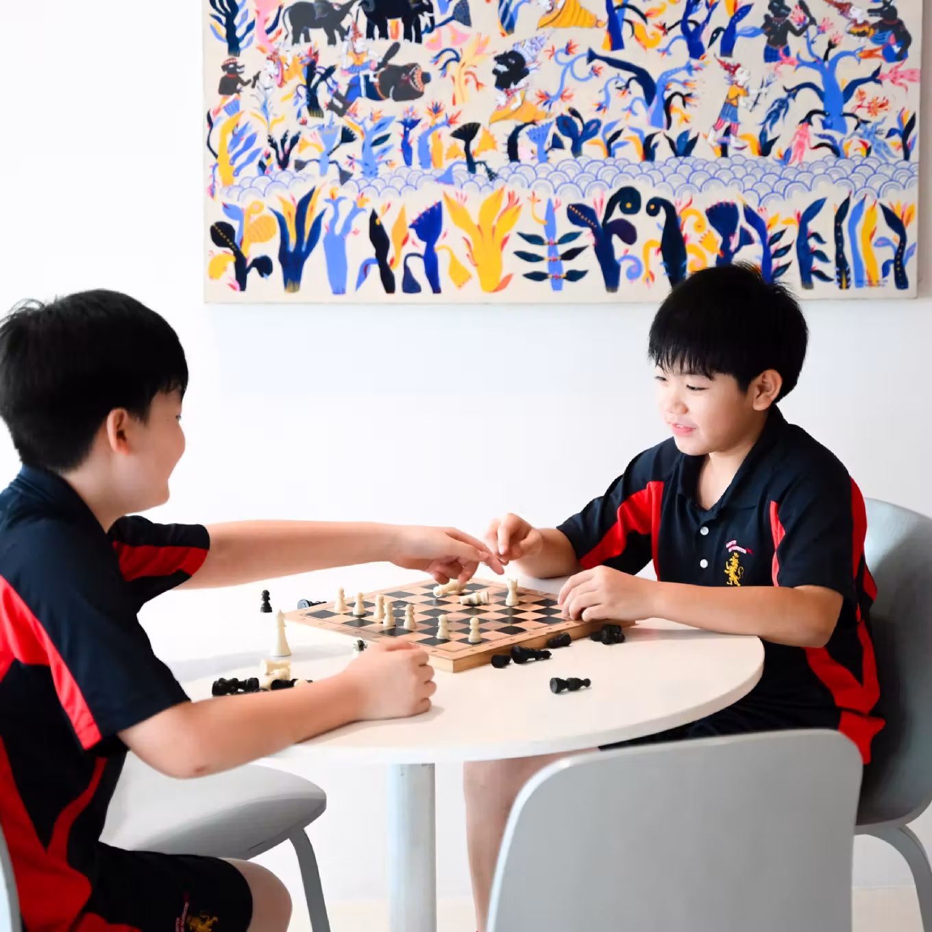 Two boys in black and red shirts playing chess at a white round table with colorful wall art behind.