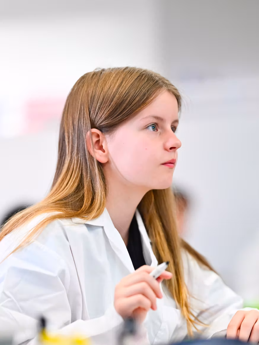 Young girl with long hair wearing a white lab coat and holding a pen, looking attentively to the side.
