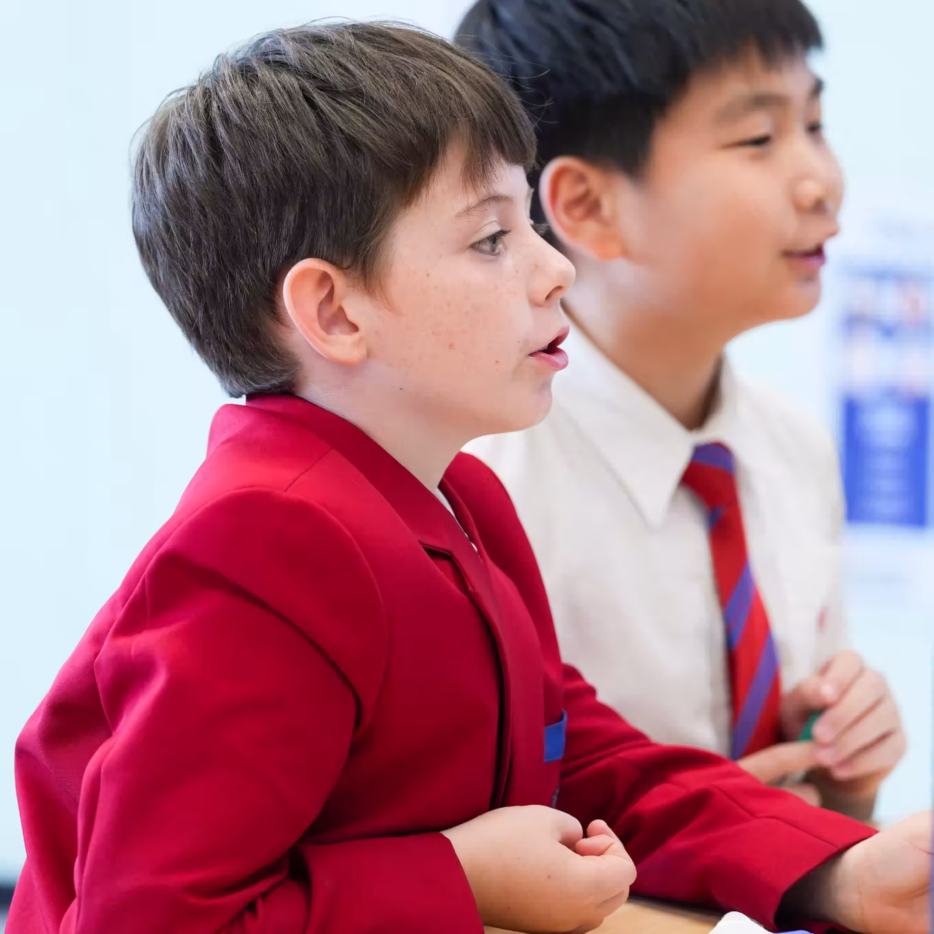 Two schoolboys in uniforms sitting at a desk, focused and possibly speaking or listening.