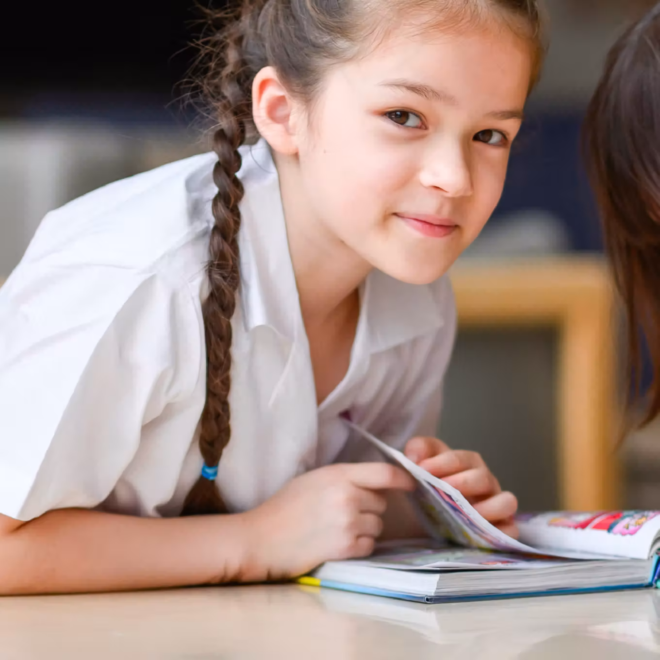 Young girl with braided hair smiling while reading a book on a table.