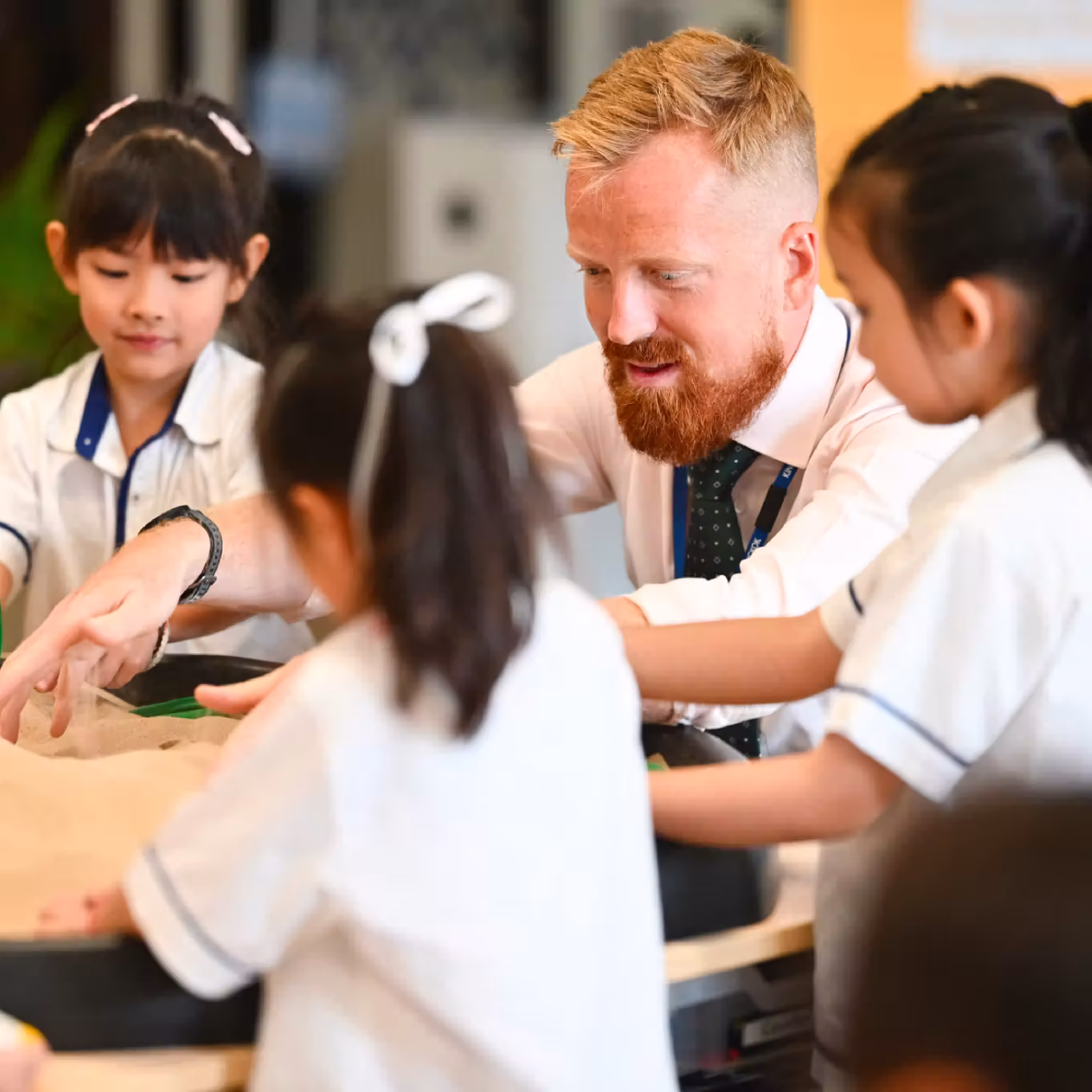 Teacher with red beard guiding three young girls in white uniforms during a hands-on learning activity.