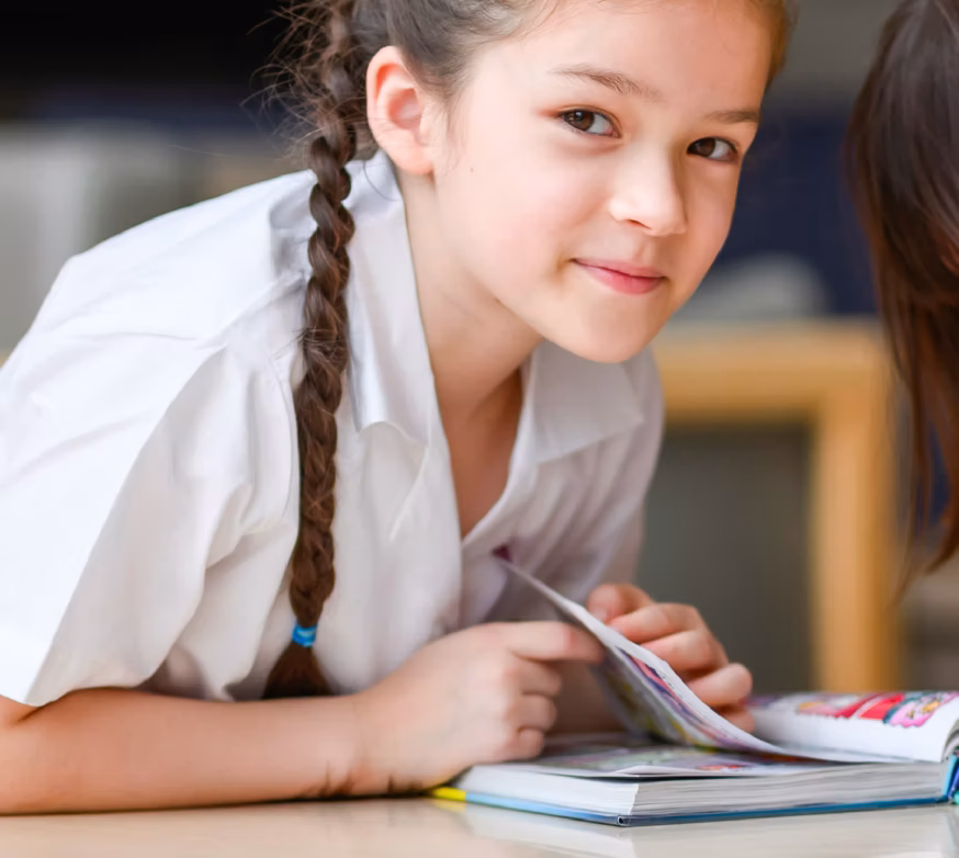 Young schoolgirl with braided hair smiling while reading a book on a table at school.