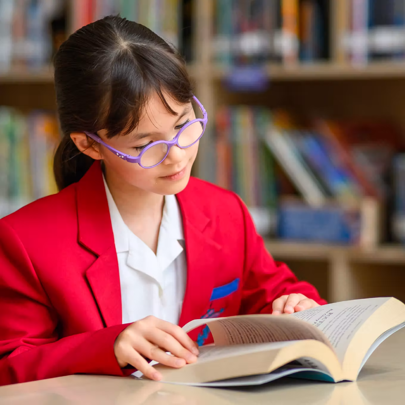 Girl wearing purple glasses and red blazer reading a book in a library with bookshelves behind her.